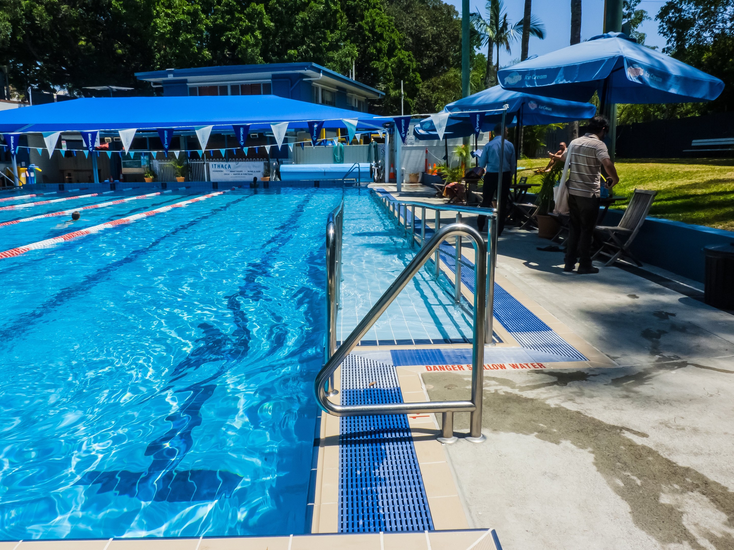 Large outdoor swimming pool with people swimming, surrounded by railings, umbrellas, chairs, tables, plants, grass, trees, and a building, under a clear blue sky.