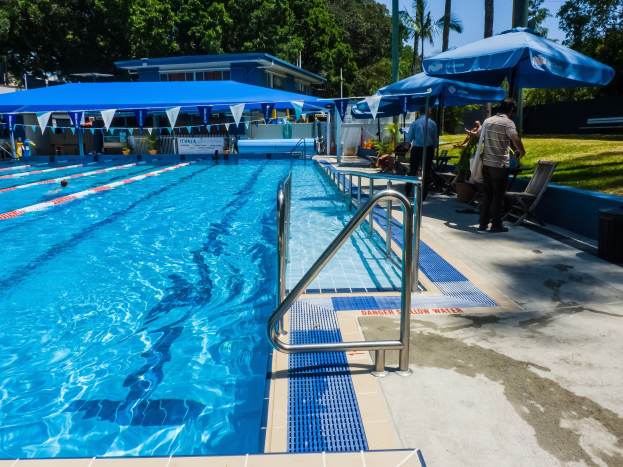 Large outdoor swimming pool with people swimming, surrounded by railings, umbrellas, chairs, tables, plants, grass, trees, and a building, under a clear blue sky.