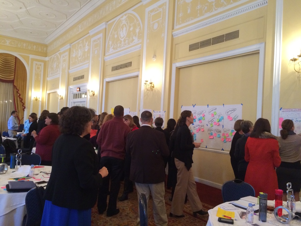 Eine Gruppe von Menschen untersucht Plakate und Charts, die an einer Wand in einem Raum zu sein scheinen, der ein Kunstmuseum zu sein scheint.