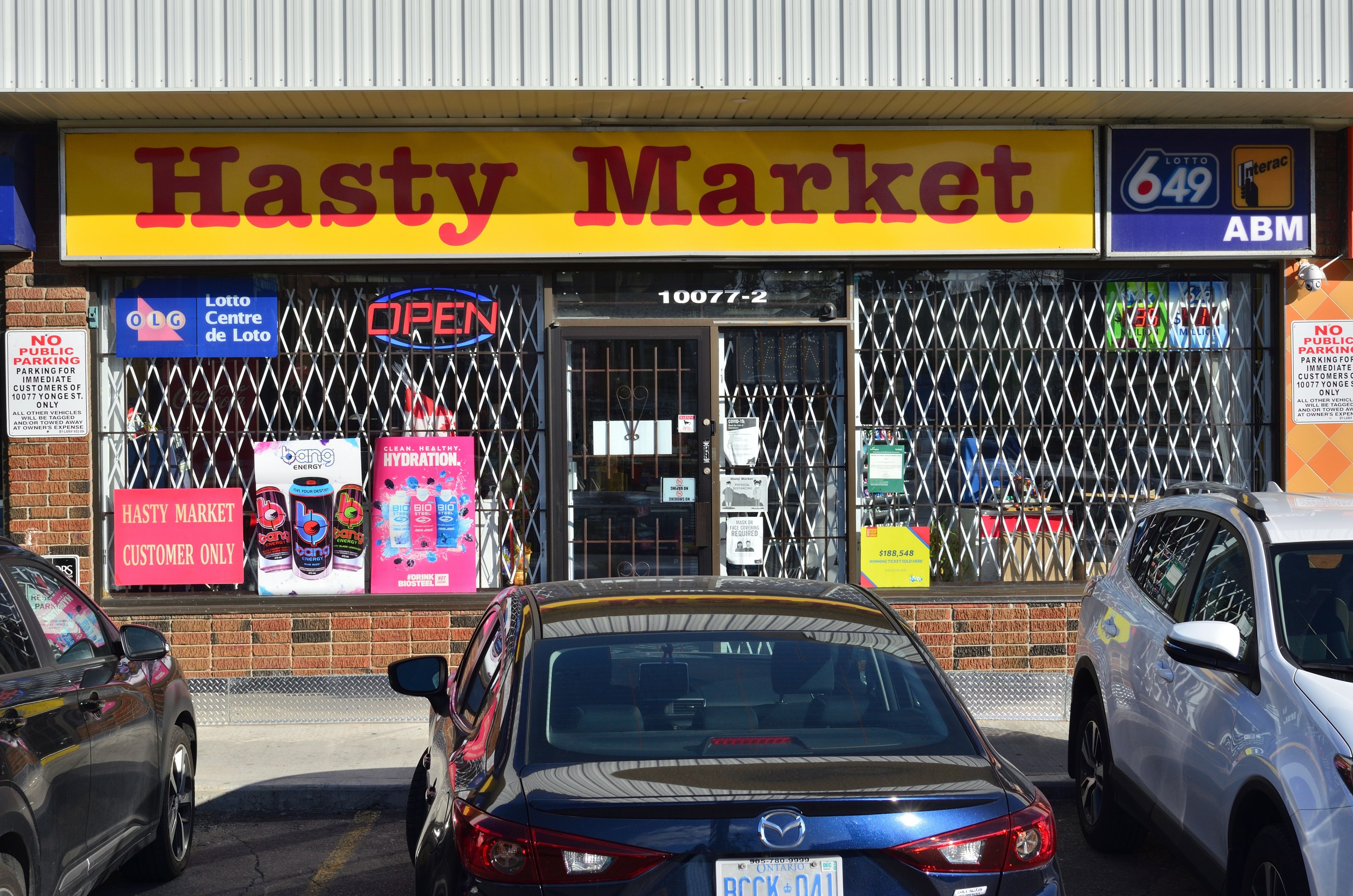 A busy Hasty Market storefront in Toronto, Ontario, with cars passing on the road in front and posters visible on the glass walls.