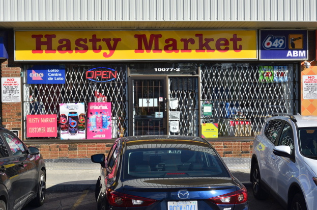 A busy Hasty Market storefront in Toronto, Ontario, with cars passing on the road in front and posters visible on the glass walls.