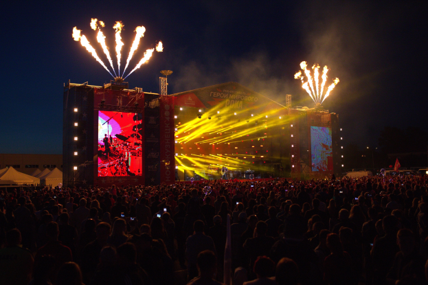 Eine große Menschenmenge auf einem Musikfestival schaut auf einer hell erleuchteten Bühne Performern zu, mit Zelten links und Feuerwerk in einem dunklen Himmel dahinter.