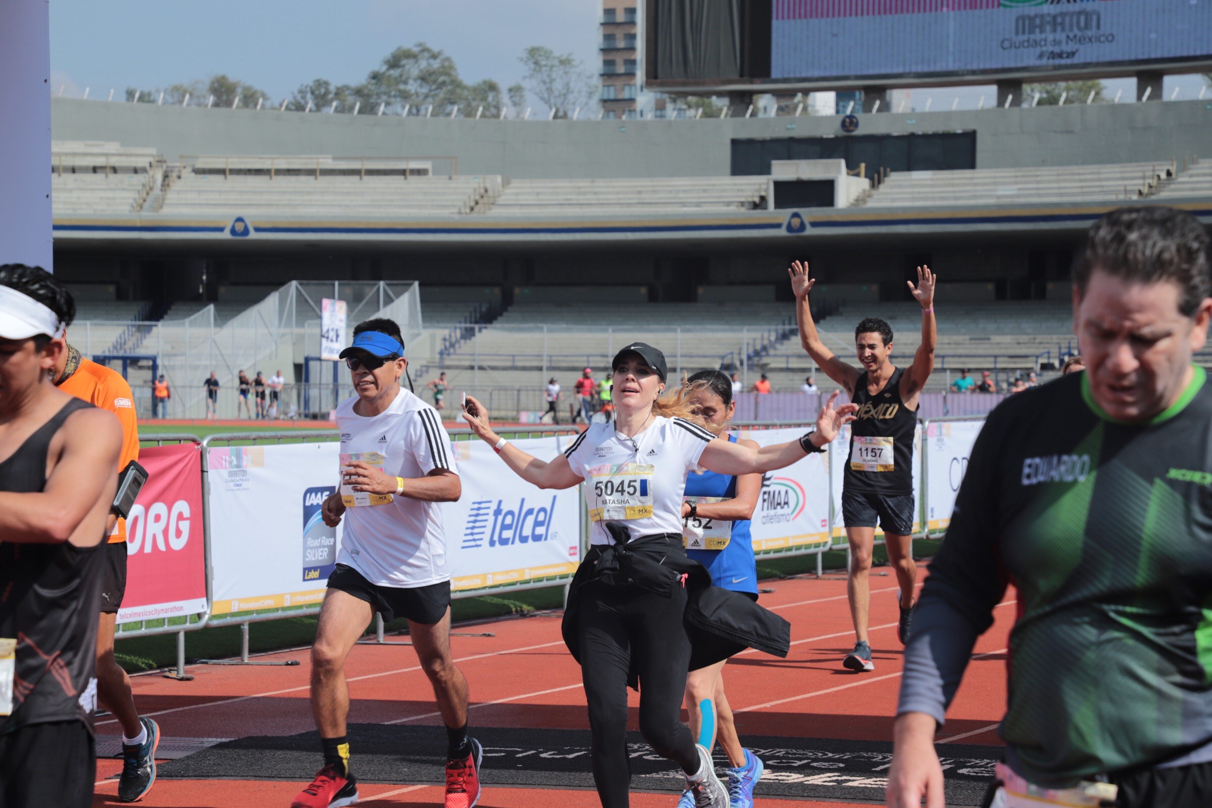 Gruppe von Menschen, die auf einer Outdoor-Sportanlage laufen, mit Bannern, Anzeigetafel, Gebäude, Bäume und bewölktem Himmel; einige Läufer tragen Turnschuhe und Hüte.