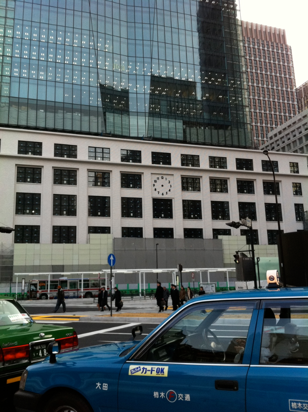 Eine Szene mit mehreren Autos, Holzplanken, einem Bus, Pfählen, Gebäuden und einigen Menschen, die stehen, mit sichtbarem Text auf einem der Autos.