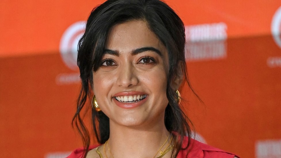 Eine Frau in einem roten Kleid mit einem eleganten Dutt und dezentem Make-up lächelt in die Kamera, trägt schwarze High Heels und hat ein hellgelbes Banner mit der Aufschrift 'Die besten Schönheits Looks von Sonam Kapoor auf dem Cannes Film Festival' im Hintergrund.