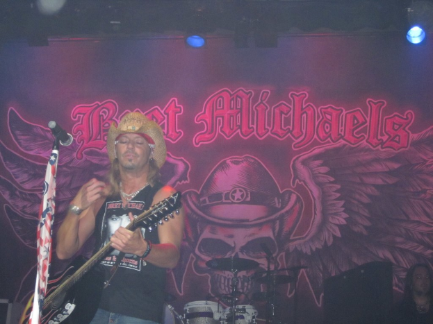 Eine musikalische Konzertsituation mit einem Gitarristen links neben einem Mikrofon, einer Person in der unteren rechten Ecke, einem Banner im Hintergrund und Drogen in der Mitte unten, alles unter Bühnenbeleuchtung von oben.