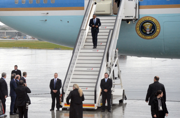 Präsident Obama besteigt Air Force One am Flughafen, begleitet von einer Gruppe in schwarzen Blazern, weißen Hemden und schwarzen Hosen, mit einem Fahrzeug, Gras, Gebäuden und einem klaren blauen Himmel im Hintergrund.