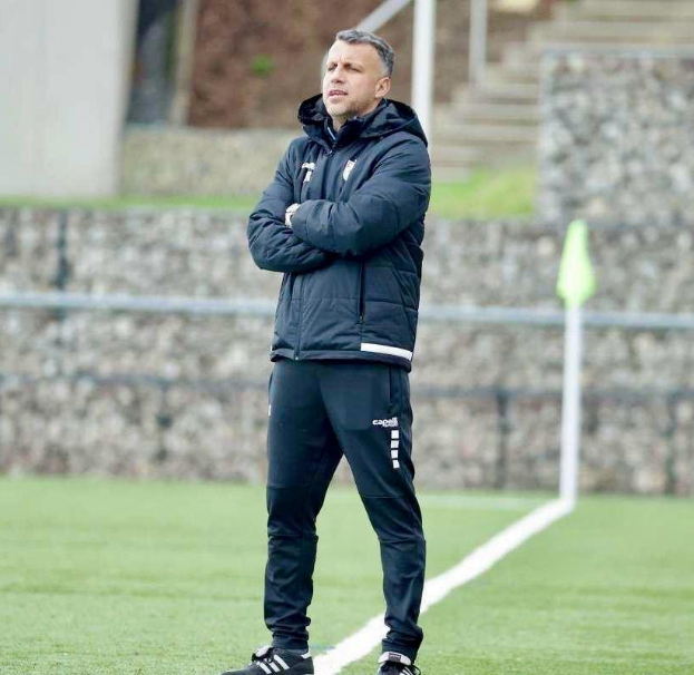 Ein Mann in schwarzer Jacke und Hose steht auf einem Fussballfeld mit überkreuzten Armen, der als Trainer während einer Trainingseinheit erscheint, mit einem Zaun, Treppe und einer Wand im Hintergrund.