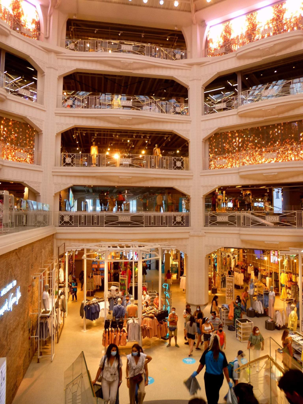 Ein belebtes Einkaufszentrum mit Menschen, die Taschen tragen und Kleider auf Schaufensterpuppen anschauen, umgeben von Geländern, Beleuchtung und Wandtafeln.