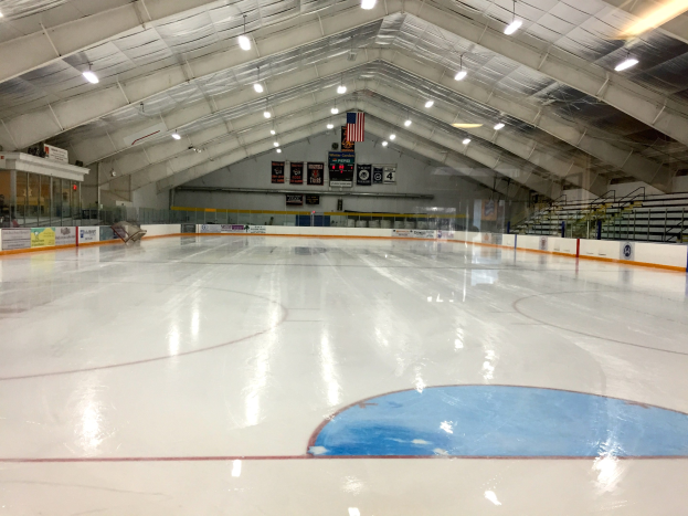 Große Indoor-Eisbahn mit Deckenbeleuchtung, Texttafeln, einer Wand, Treppen mit Geländern und einigen Menschen, die einem Hockey-Sportplatz ähnelt.