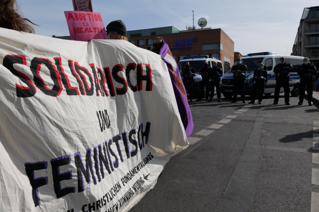 Eine Gruppe von Menschen marschiert auf einer Straße, hält ein Banner mit der Aufschrift 'Solidarität und Feminismus', mit parkenden Fahrzeugen am Straßenrand und Gebäuden im Hintergrund unter einem klaren blauen Himmel.