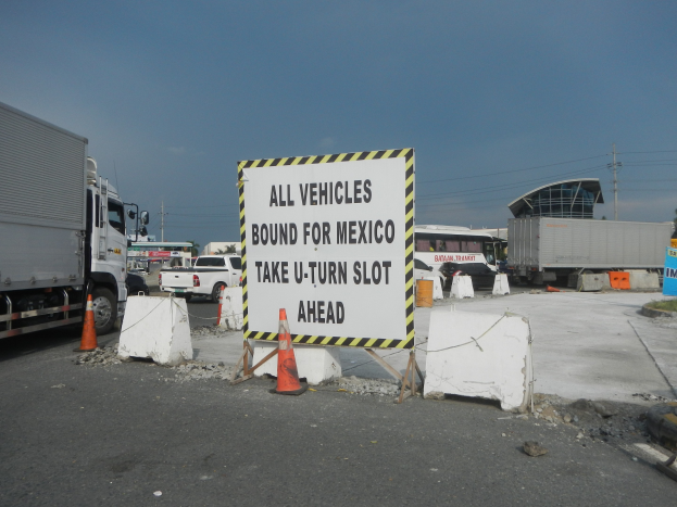Lastwagen auf einem Parkplatz neben einem Schild "Alle Fahrzeuge nach Mexiko" mit verstreuten Verkehrskegeln, Brettern und Hintergrundgebäuden, Bäumen, Polen und einem klaren blauen Himmel.