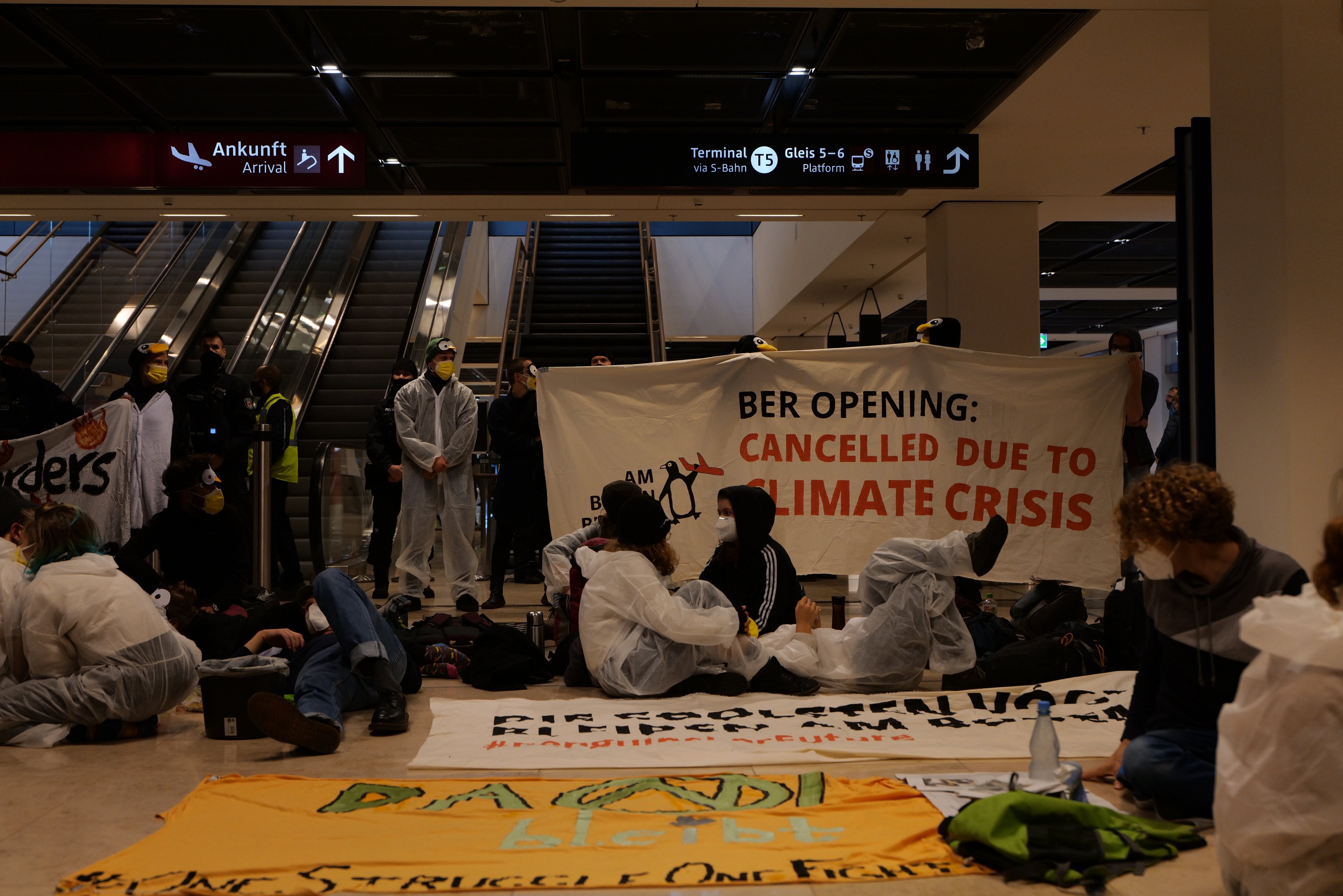 A group of people sitting on an airport floor holding a "Berlin Cancelled Due to Climate Crisis" banner, with scattered bags, bottles, and background escalators, text boards, and ceiling lights.