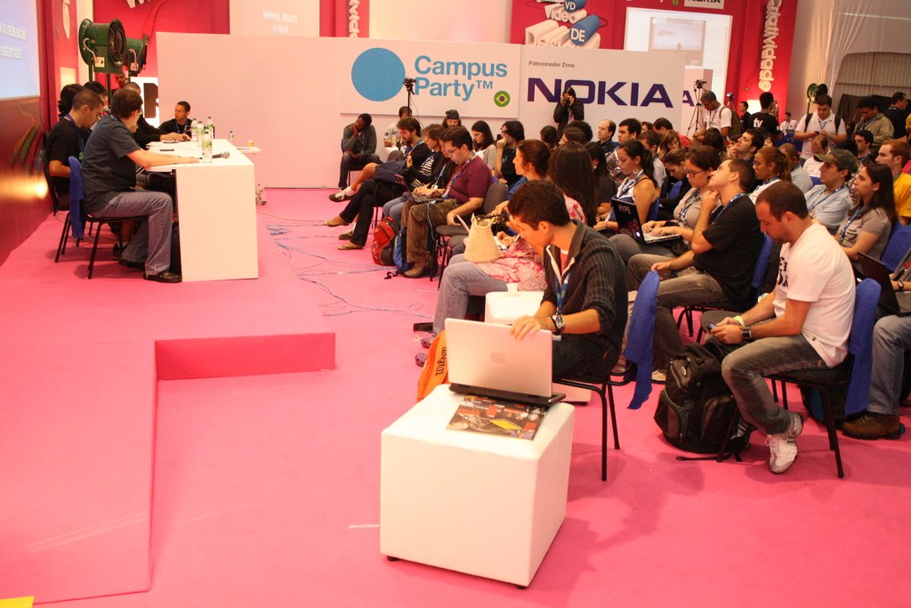 Conference with seated attendees listening to speakers on stage, media and owners conversing, with a large hoarding and wall in the background.