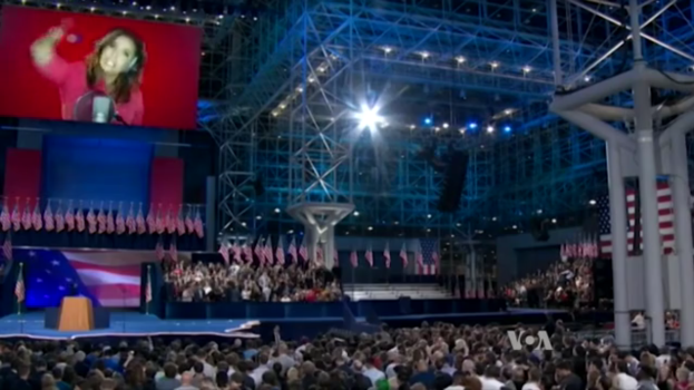 Eine große Menschenmenge sitzt vor einer Bühne mit einem Rednerpult, Mikrofon, Flaggen, einem Bildschirm mit dem Bild einer Frau und Lichtausrüstung, wahrscheinlich bei einer Präsidentschaftsdebatte.