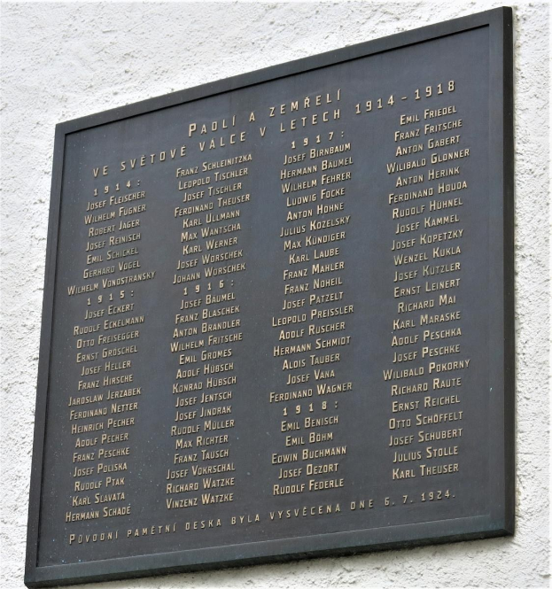 A stone plaque with inscribed text, likely a Holocaust memorial, mounted on a stone wall with plants at its base.