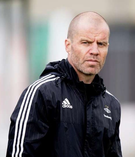 Ein kahlköpfiger Trainer in einer blauen Jacke mit weißen Streifen und weißem Kragen steht auf einem Fußballfeld, die Hände verschränkt und mit einem ernsten Gesichtsausdruck.