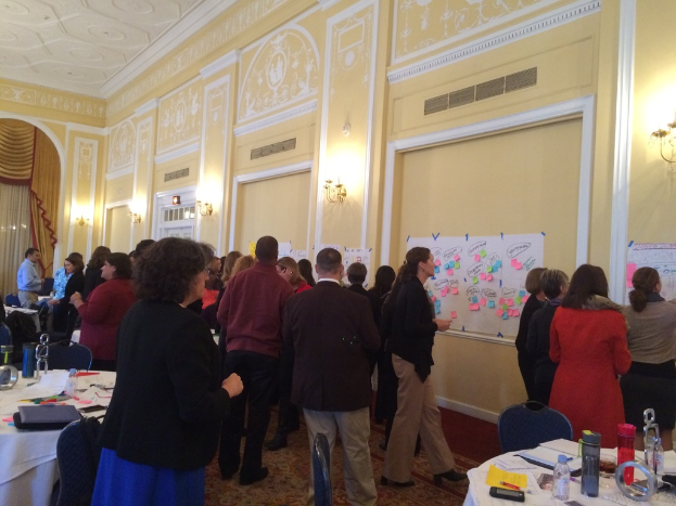 Eine Gruppe von Menschen betrachtet Plakate und Charts an einer Wand in einem scheinbar künstlerischen Museum.