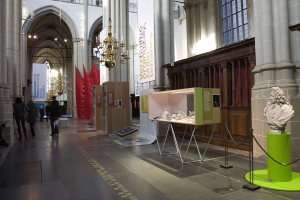 A large museum room displaying various artifacts, including a statue on a pedestal, roped stands, banners with text, pillars, and chandeliers, with a few people present.