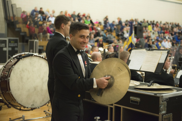 Eine Gruppe von Männern in Fracks, die als Band auf der Bühne spielen, wobei einer eine Becken hält und lächelt, während andere Instrumente spielen, mit Büchern auf Ständern und einer Wand im Hintergrund.