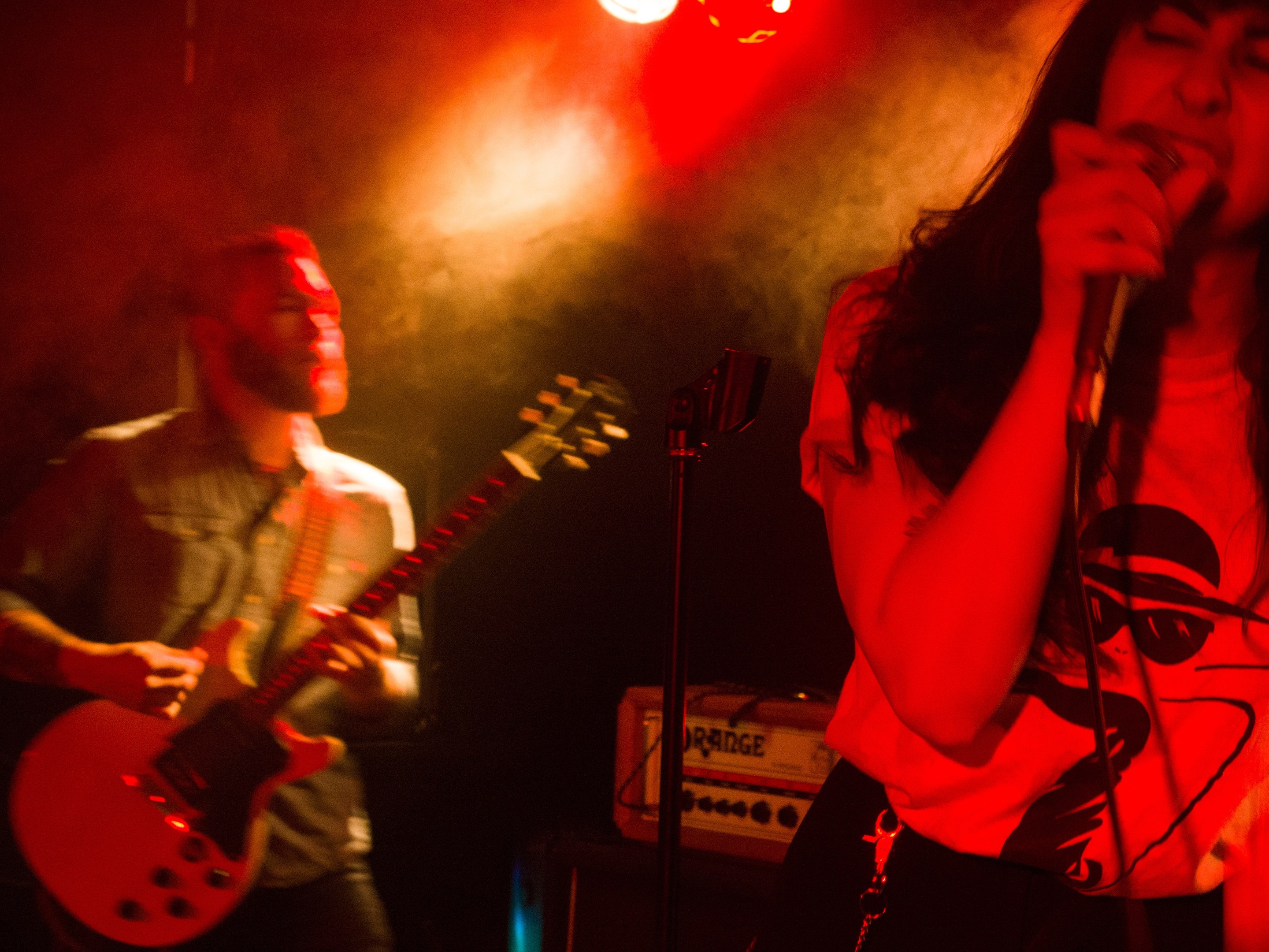 Eine Frau singt in ein Mikrofon auf der Bühne, während ein Mann im Hintergrund eine akustische Gitarre spielt und Deckenlampen das lebhafte Setting beleuchten.