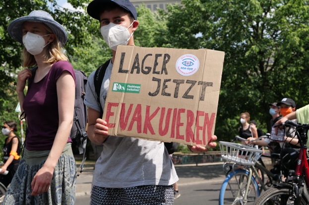 Ein Mann und eine Frau mit Masken halten ein Schild mit der Aufschrift "Lager Jetzt Evakuieren" vor einer Gruppe mit Fahrrädern, Bäumen und einem Gebäude im Hintergrund.