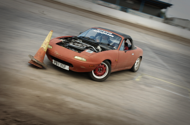 Eine Person fährt ein rotes Auto mit hoher Geschwindigkeit auf einer Rennstrecke und nähert sich einem Verkehrskegel.