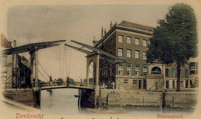 Old black-and-white photograph of a bridge under construction spanning a river, with buildings, trees, and a clear sky in the background and scattered tools and materials along its base.