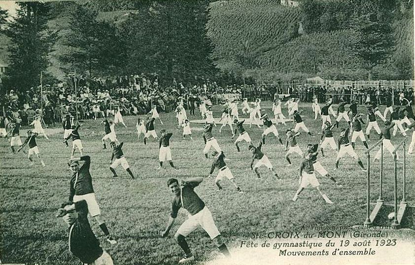 Black and white photo of a group of people playing soccer on a grassy field with trees, buildings, and a clear sky in the background, dated "Fête de Gymnastique du 19 août 1923".