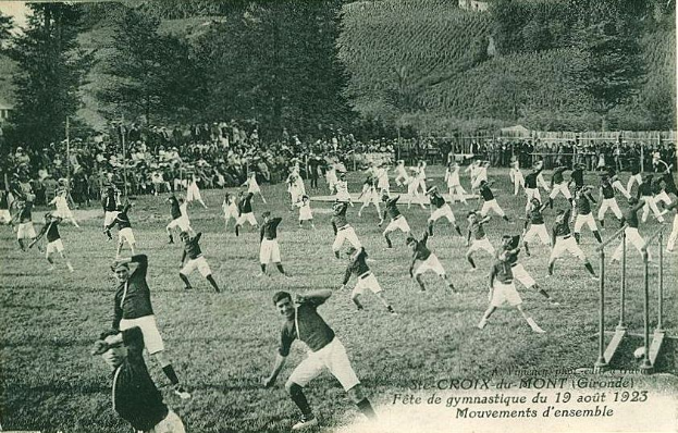 Black and white photo of a group of people playing soccer on a grassy field with trees, buildings, and a clear sky in the background, dated "Fête de Gymnastique du 19 août 1923".