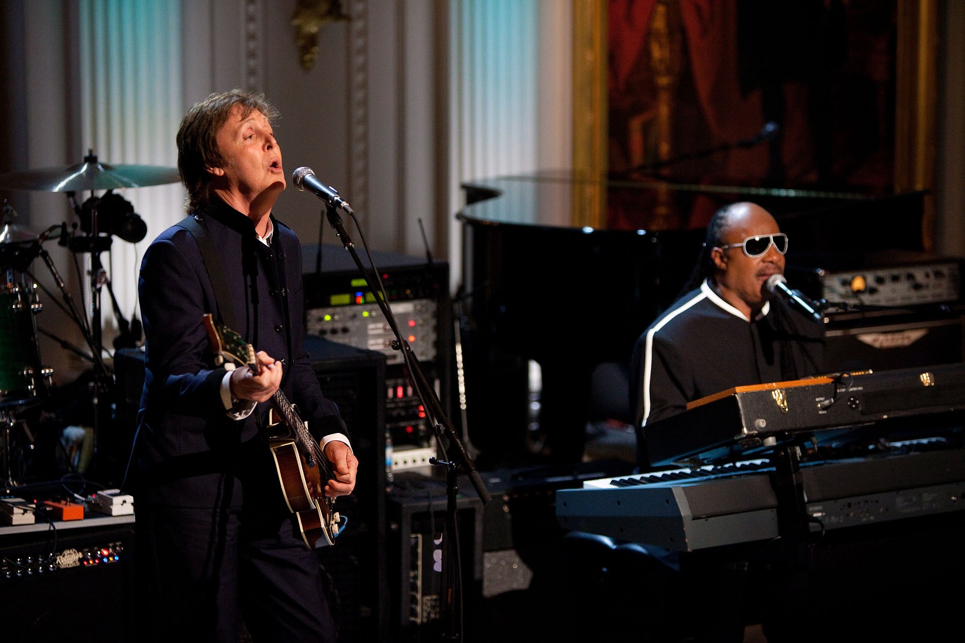 Paul McCartney spielt Gitarre und singt in ein Mikrofon, während Stevie Wonder eine Piano spielt und eine Brille trägt auf der Bühne bei den Grammys, mit Musikinstrumenten und gerahmten Kunstwerken im Hintergrund.