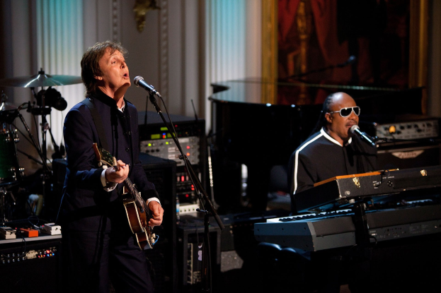 Paul McCartney spielt Gitarre und singt in ein Mikrofon, während Stevie Wonder eine Piano spielt und eine Brille trägt auf der Bühne bei den Grammys, mit Musikinstrumenten und gerahmten Kunstwerken im Hintergrund.