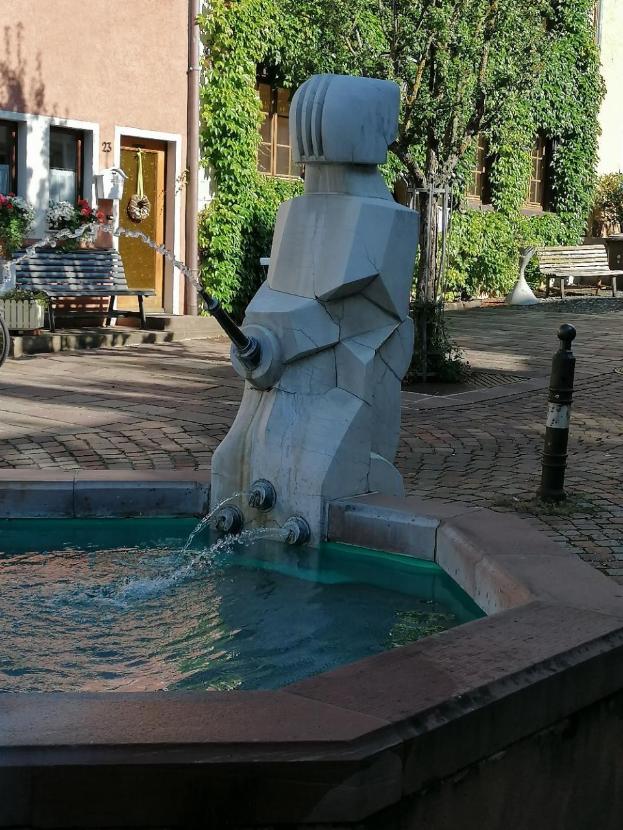 Ein zentraler Stadtplatzbrunnen umgeben von Gebäuden, Grünflächen, Bänken und Blumenkübeln, mit Wasser im Becken.