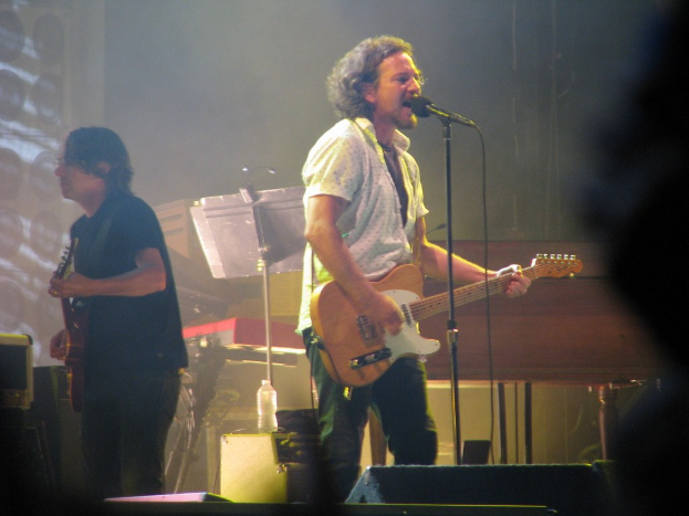 A live music performance on stage with a man in a white shirt singing into a microphone and playing guitar, accompanied by another man in a black shirt playing guitar; a piano, cables, speakers, and stage lights are visible in the background.