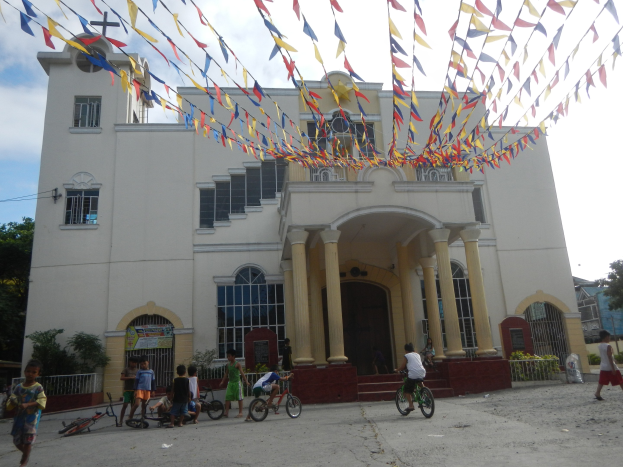 Gruppe von Menschen, die Fahrräder vor einer weißen Kirche mit architektonischen Details fahren, umgeben von Grün, einem Banner, Bäumen und bunten Fahnen vor einem bewölkten Himmel.