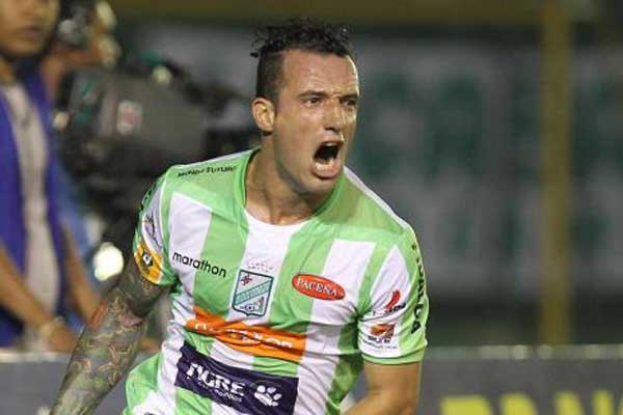 Ein Fußballspieler in grün-weißer Uniform mit Tattoos auf dem Arm feiert nach einem Tor, mit einem unscharfen Hintergrund von Menschen und einer Tafel mit unleserlicher Schrift.