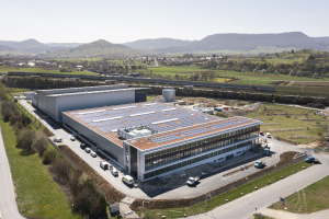 Aerial view of a large factory building with solar panels on its roof, surrounded by vehicles, trees, grass, and utility poles, with hills and a clear blue sky in the background.