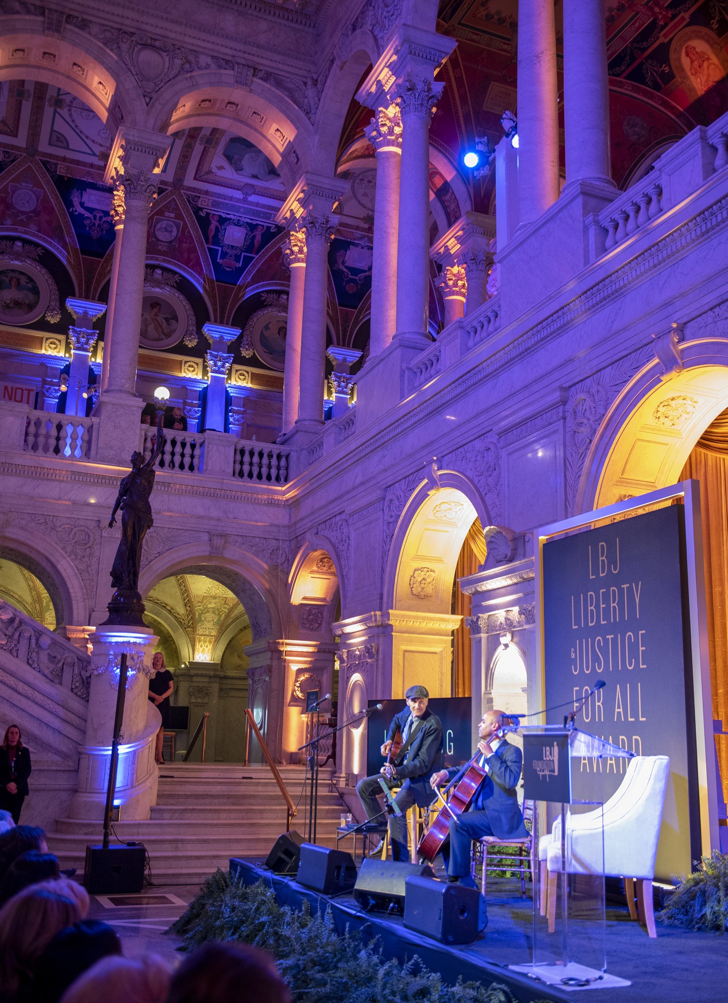 A group of people seated on a stage during the Liberty Justice for All Award Ceremony in New York City, with musicians, a podium, a microphone, a board with text, and architectural elements including a statue, railings, pillars, and ceiling lights.