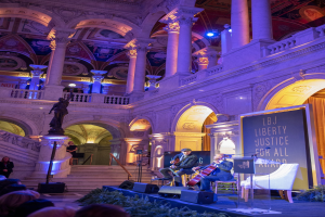 A group of people seated on a stage during the Liberty Justice for All Award Ceremony in New York City, with musicians, a podium, a microphone, a board with text, and architectural elements including a statue, railings, pillars, and ceiling lights.