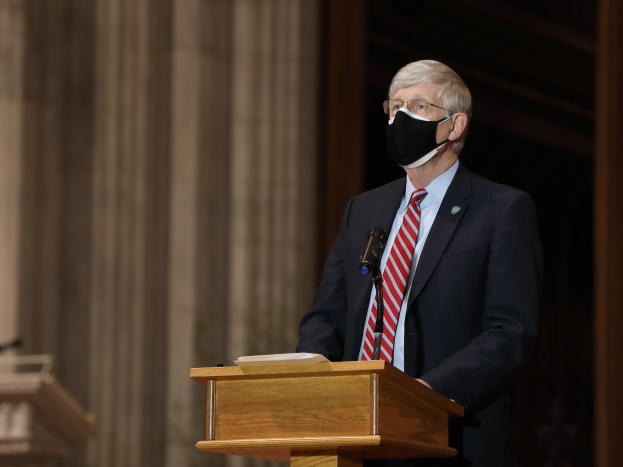 Ein Mann im Anzug und Krawatte mit einer Gesichtsmaske steht an einem Podium mit einem Mikrofon und einem Buch und hält eine Rede mit Säulen und einer Wand dahinter und einem weiteren Podium links von ihm.