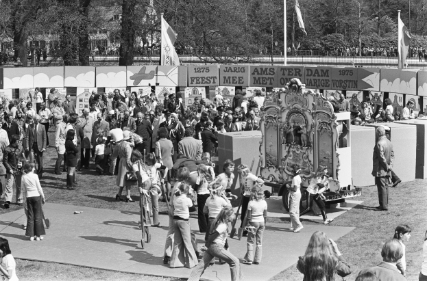 Ein Schwarz-Weiß-Bild einer Menge, die sich um einen Faschingswagen schart, der mit Fahnen und Transparenten geschmückt ist, vor einem Hintergrund aus Bäumen und Gebäuden.
