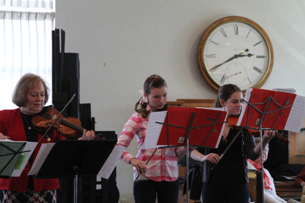Drei Frauen, die auf der Bühne Geigen spielen, mit Notenpulten und Noten davor, während ein Zuschauer im Vordergrund sitzt und eine Uhr an der weißen Wand im Hintergrund hängt, mit einem weißen Vorhang in der linken Ecke.