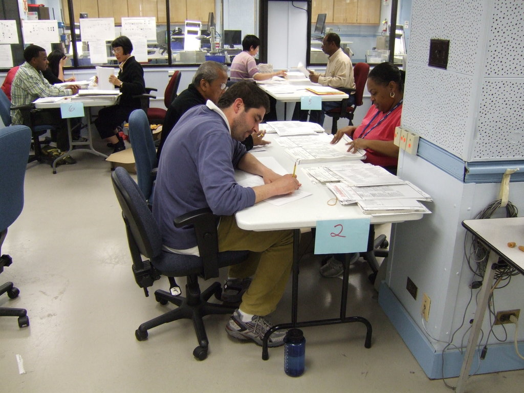 Mitarbeiter sitzen an Schreibtischen mit Papieren, einige schreiben und andere erklären, mit Glasfenstern und Monitoren im Hintergrund.