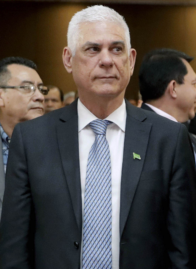 Ein Mann in einem marineblauen Anzug, weißem Hemd und gemustertem Schlips steht vor einer Gruppe von Menschen und spricht mit ernster Miene.