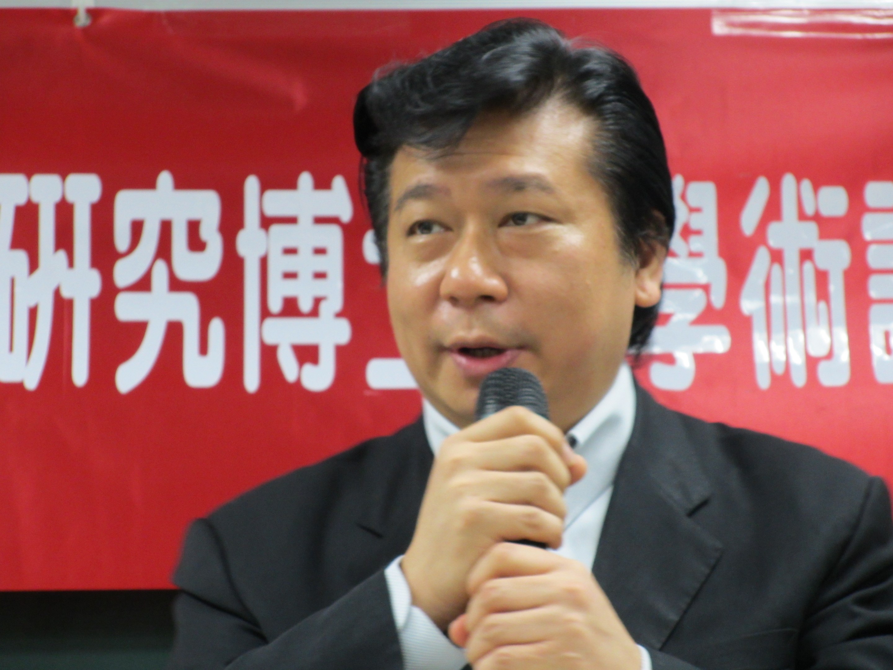 Ein förmlich gekleideter Mann in einem Anzug spricht in ein Mikrofon an einem Podium mit einem ernsten Gesichtsausdruck, vor einer Wandbanner.