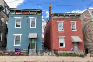 Colorful row of houses with windows, doors, a metal fence, a rental signboard, potted plants, a trash bin, wires, and a cloudy sky.