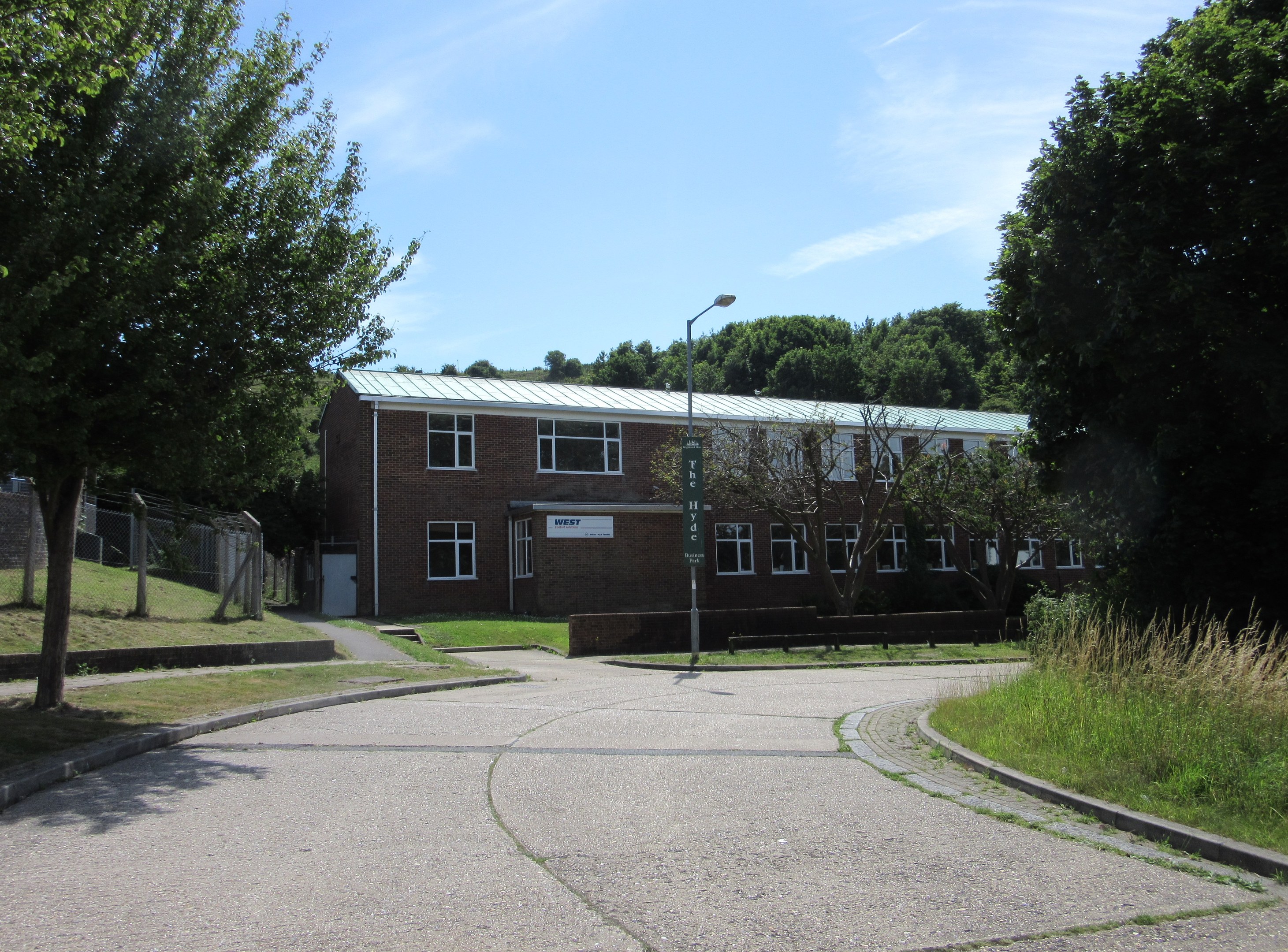 A school building with windows, a street pole, a signboard, a group of trees, grass, a fence, and a cloudy sky, situated in the middle of a park surrounded by lush green grass.