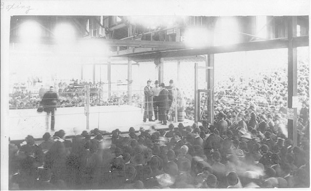 Schwarzes und weißes Foto einer Menge in einer Arena, die einen Boxkampf beobachten, mit einigen Menschen auf der Bühne und anderen im Publikum.