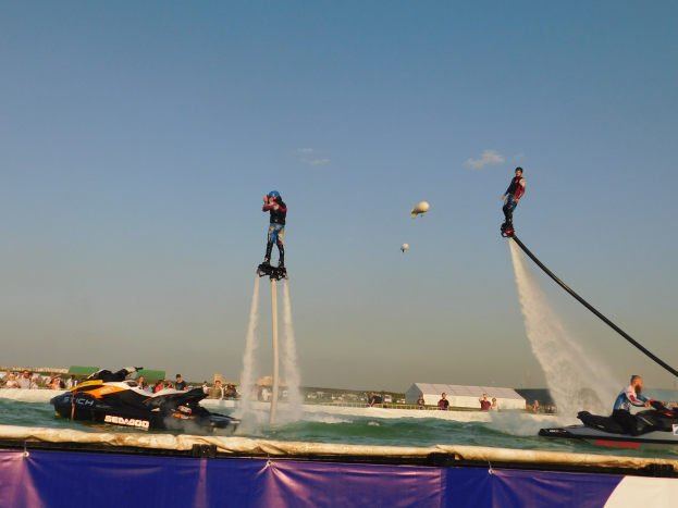 Group of people riding jet skis on water, with a banner below, against a backdrop of people, buildings, and a clear blue sky.