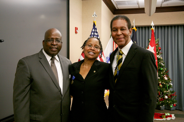Eine Gruppe strahlender Menschen vor einem geschmückten Weihnachtsbaum mit verschiedenen Gegenständen, Flaggen und einem Vorhang im Hintergrund, mit einem Tuch und zusätzlichen Gegenständen auf der rechten Seite.